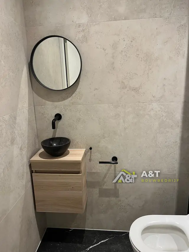 Toilet met zwarte fontein. Lichte betonlook-tegels, zwarte waskom op een houten kastje en een ronde spiegel.